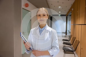 Attractive blonde young female doctor standing in hospital corridor and holding clipboard.