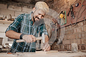 Attractive blonde young hipster bearded man by profession carpenter builder nailing wood board with hammer on a wooden