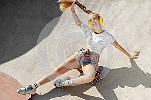 Attractive blonde young woman sitting on yellow skateboard or pennyboard and listening music at the skatepark. Freedom