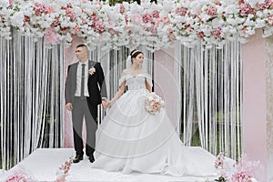 Attractive bride and groom at the ceremony on their wedding day with an arch made of pink and white flowers