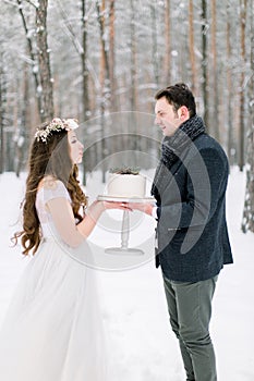 Attractive bride with long curly hair standing together with her groom in stylish jacket, holding wedding cake in the