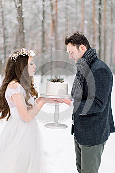 Attractive bride with long curly hair standing together with her groom in stylish jacket, holding wedding cake in the