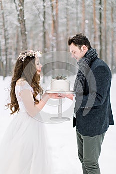 Attractive bride with long curly hair standing together with her groom in stylish jacket, holding wedding cake in the