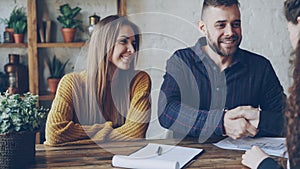 Attractive broker is selling house to young couple, people are signing documents and shaking hands, realtor is giving