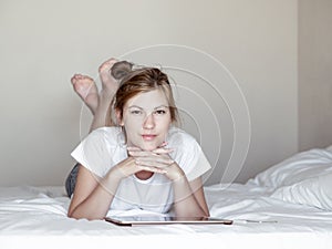 Attractive brown-haired girl lying on the bed next to the tablet and phone and smiling at camera in bedroom