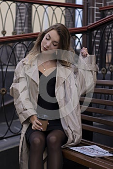 Attractive brunette in fashionable trench coat and black dress sitting on bench with a newspaper. autumn, yellow leaves