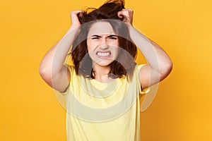 Attractive brunette female scrathes head and exclaims loudly, looks angry, wears casual t shirt, poses against yellow concrete