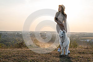 Attractive brunette girl in beige dress and hat stands among the field with samoed dog, beautiful landscape on the