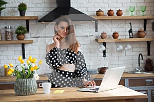 Attractive brunette girl blogger in a polka dot dress sitting in the cozy kitchen and reading news on laptop at the wooden table