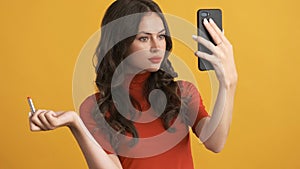 Attractive brunette girl confidently applying red lipstick looking in smartphone over yellow background