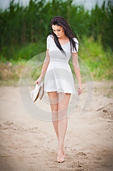 Attractive brunette girl with short white dress strolling barefoot on the countryside road. Young beautiful woman walking