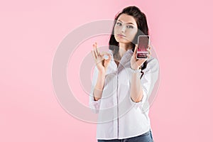 Attractive brunette girl showing ok sign and smartphone with trading courses on screen, isolated