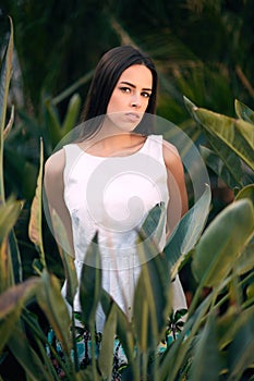 An attractive brunette girl. A thoughtful young female. A lady in a white dress on a tropical garden background. A