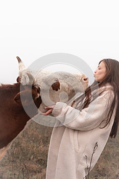 Attractive brunette girl in white coat blows a kiss to a cow, meadow in fog on the background