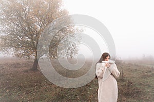 Attractive brunette girl wrapped in white coat stands among the meadow, big tree in fog on the background