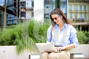 Attractive brunette haired woman on a bench in the city using laptop for work