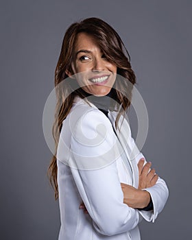 Attractive brunette haired woman wearing white blazer while standing at isolated dark grey background