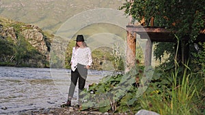 Attractive brunette in a hat and a white shirt in peas enjoys nature in the mountains. Young woman tourist walks along