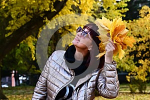 an attractive brunette holds bouquet of yellow maple leaves. Lady enjoys autumn nature and smiles carelessly.