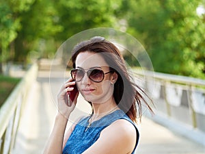 Attractive brunette woman in sunglasses and blue jeans dress has emotional phone conversation on mobile phone on park bridge outdo