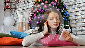 Attractive brunette lying on bright pillows near the Christmas tree and listening to music. Unconventional Christmas