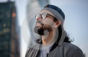 Attractive brunette man in cap wearing glasses in big city, head profile shot, street style