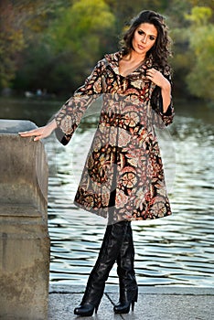 Attractive brunette model wearing elegant coat posing next to lake in the autumn park.