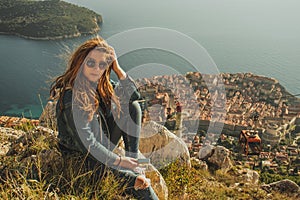 Attractive brunette sitting on the Srd hill above the city of Dubrovnik, observing the city in the distance. Wind blowing her hair