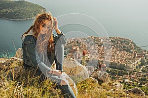 Attractive brunette sitting on the Srd hill above the city of Dubrovnik, observing the city in the distance. Wind blowing her hair
