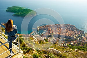 Attractive brunette traveller sitting on a railing above the city of Dubrovnik, viewpoint on Srd mountain. Looking at the ancient