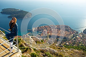 Attractive brunette traveller sitting on a railing above the city of Dubrovnik, viewpoint on Srd mountain. Looking at the ancient