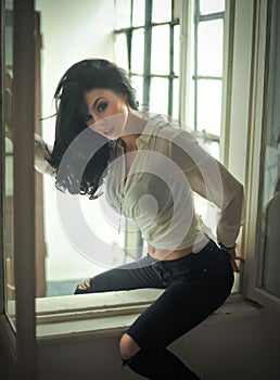 Attractive brunette in white tight fit shirt and black ripped jeans posing sitting on window frame. Sensual modern woman