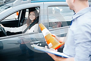 Attractive brunette woman on driver`s seat with fastened seat belt smiles to male driving instructor with clipboard and