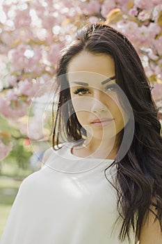 Attractive brunette woman in light dress looking at camera while posing near flowering tree