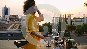 Attractive brunette woman in a long yellow dress during the dawn standing by her city bicycle holding its handlebar with