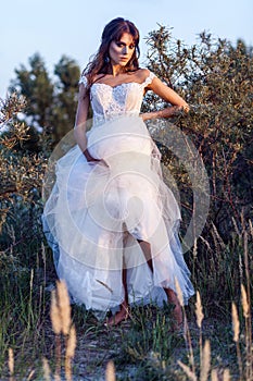 Attractive brunette woman with makeup and hairstyle wearing white wedding dress while posing at glade. holding dress and looking