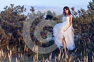 Attractive brunette woman with makeup and hairstyle wearing white wedding dress while posing at glade. holding her dress and