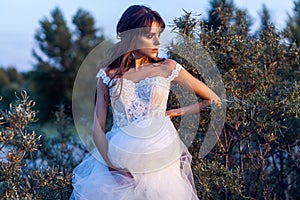 Attractive brunette woman with makeup and hairstyle wearing white wedding dress while posing at glade. looking away with hand on
