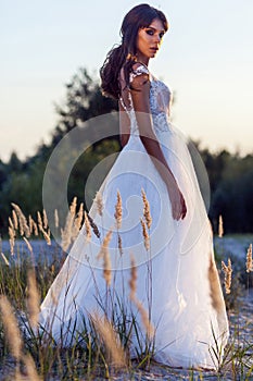 Attractive brunette woman with makeup and hairstyle wearing white wedding dress while posing at glade. side view and looking at