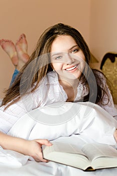 Attractive brunette young woman beautiful happy smiling student girl in bed with book looking at camera portrait