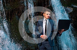 Attractive business man in suit holding laptop at waterfall background. Telework stressful work concept.