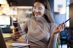 Attractive business woman sits at table in front of laptop, holds smartphone and pencil in her hand.