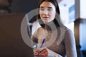 Attractive business woman sits at table in front of laptop, makes notes in notebook, draws up business schedule.