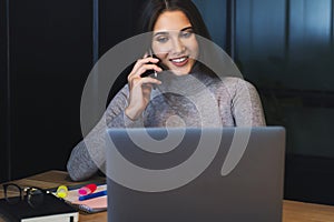 Attractive business woman sits at table in front of laptop and talks on mobile phone