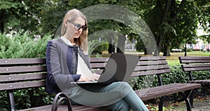 attractive business woman with sunglasses sitting on the bench in park and working on laptop