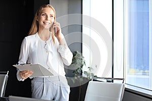 Attractive business woman talking with collegues on the mobile phone while standing near the window in the office