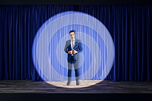 Attractive businessman standing in front of blue curtains in spotlight in theater. Presentation and speech concept