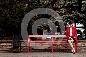 Attractive businesswoman is doing notes in paper notebook on bench in the park. Executive in red blazer and lether skirt