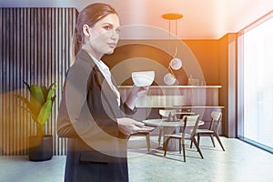 Attractive businesswoman in formal suit is drinking coffee at corporate office kitchen. Modern kitchen set and dining area as