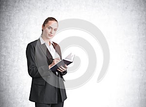 Attractive businesswoman in formal suit and shirt is holding a planner and pen, taking notes, looking straight camera and dreaming